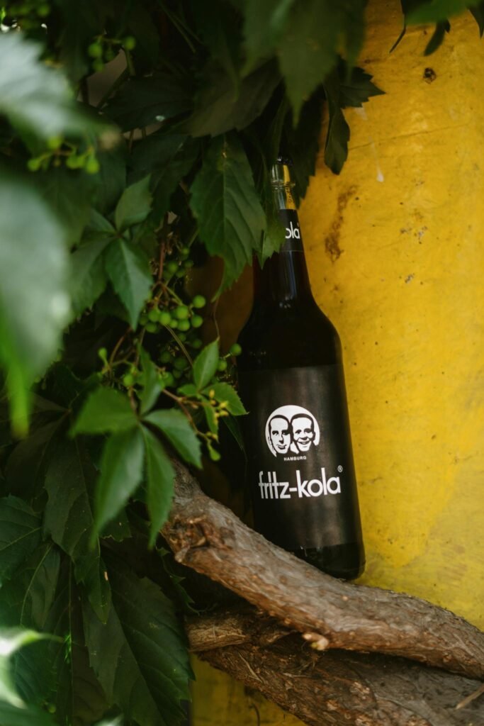 A Fritz-Kola bottle nestled amongst green leaves and branches against a vibrant yellow backdrop.